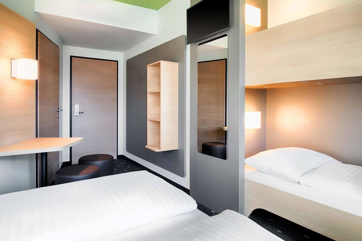 A modern hotel room with a bunk bed, a bedside table, a mirror, and a small seating area with stools.