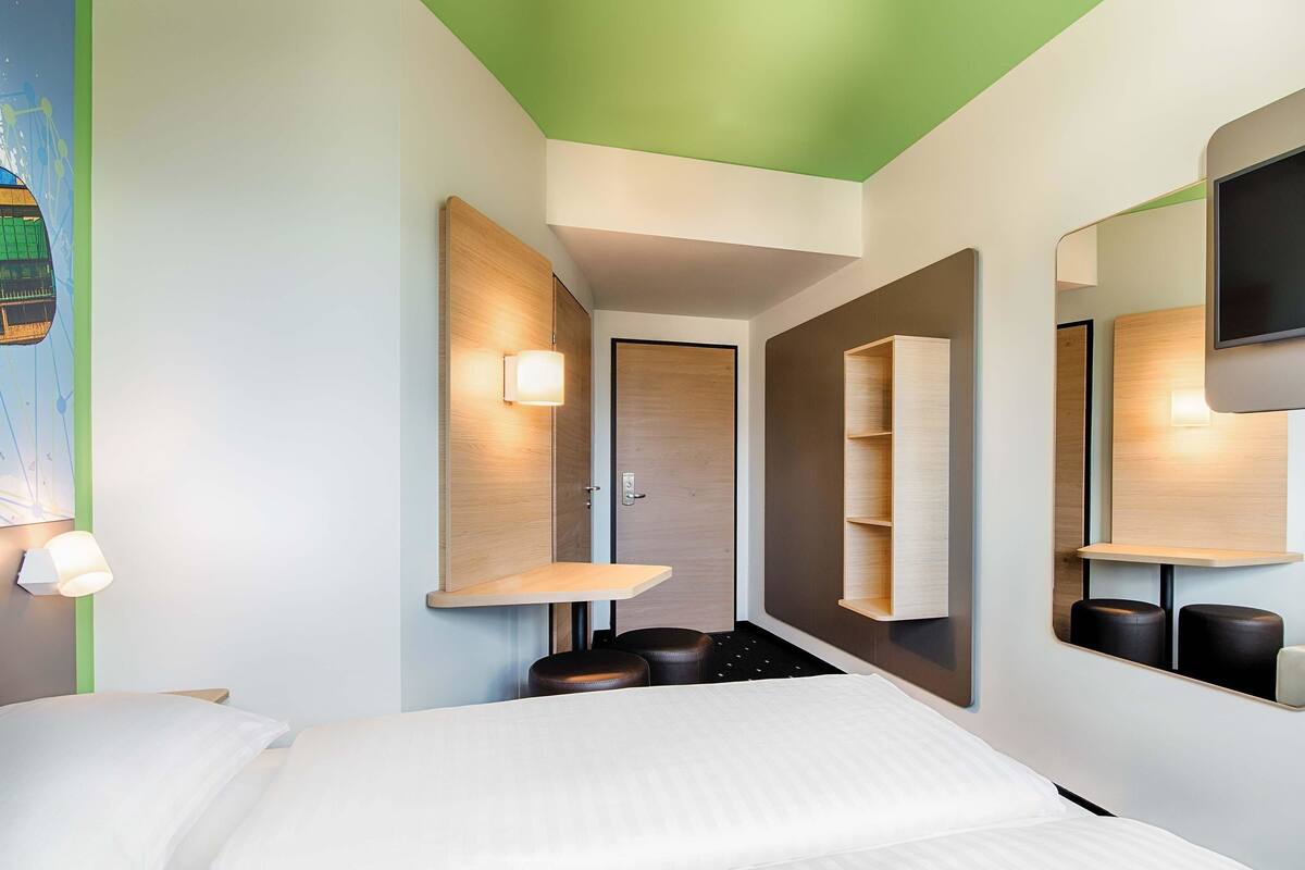 A hotel room with a bed, a nightstand, a mirror, and a wall-mounted shelf.