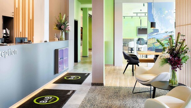 Zona de recepción con un mostrador de madera, pared verde y un área de estar con mesa y sillas.