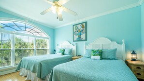 Quarto com duas camas, um ventilador de teto, uma janela com vista para área verde e um quadro na parede.