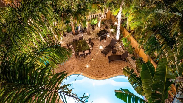 Área de piscina ao ar livre com um pátio de tijolos, cadeiras de descanso e uma mesa de jantar, tudo cercado por vegetação exuberante e palmeiras.
