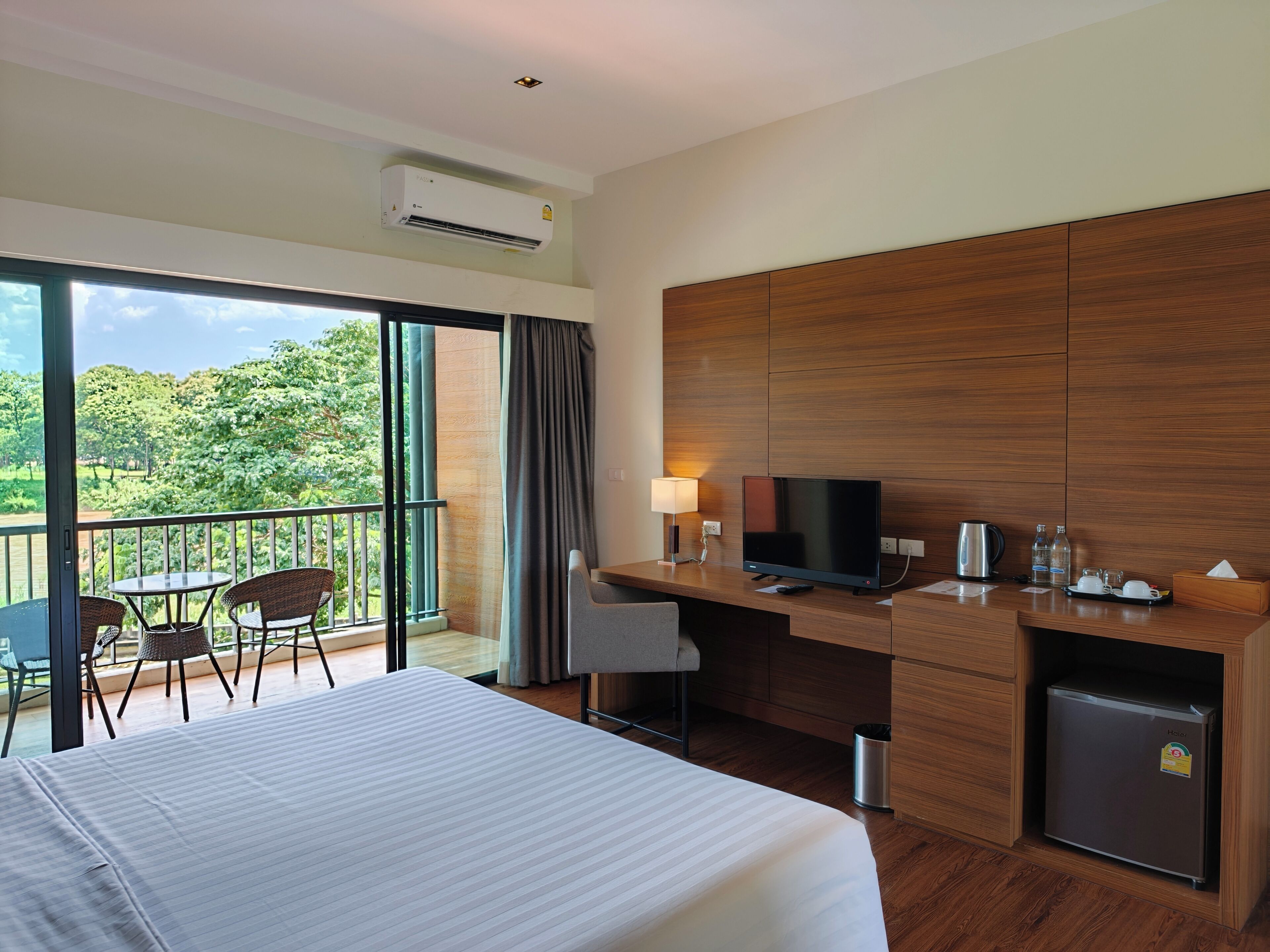 Ein Hotelzimmer mit einem Bett, einem Schreibtisch mit Fernseher, einer Minibar, einem Mülleimer und einem Balkon mit Stühlen und Tisch.
