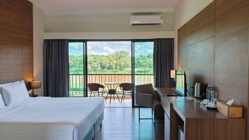A hotel room with a bed, a desk, chairs, and a balcony with a view of greenery.