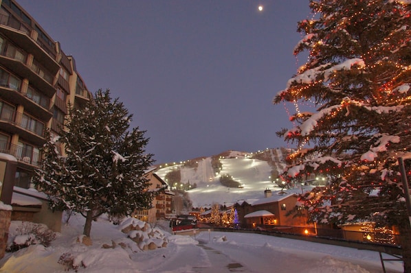 A snow-covered landscape with a ski slope, illuminated by lights, and a building with balconies.