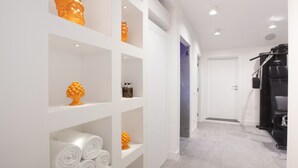 Corredor com estantes brancas exibindo objetos decorativos e toalhas, que leva a uma porta e a uma área de ginástica.