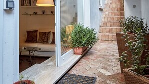 Um corredor externo estreito com uma porta de vidro deslizante que dá para uma sala de estar, uma planta em vaso e uma pequena mesa.