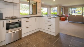 A modern kitchen with white cabinets, stainless steel appliances, and a large window offering a view of greenery. - Villa 51 - Pauanui Holiday Home (Pauanui)