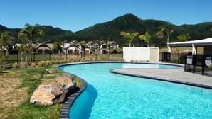 A swimming pool with a curved edge, surrounded by a fence, with houses and a mountain in the background. - Villa 51 - Pauanui Holiday Home (Pauanui)