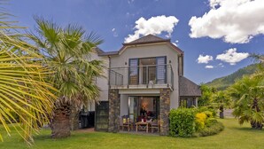 A two-story house with a balcony, surrounded by a well-maintained lawn and palm trees. - Villa 51 - Pauanui Holiday Home (Pauanui)