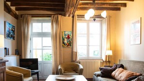 A cozy living room with a sofa, armchair, and table. There are two windows with curtains, a radiator, and a painting on the wall.