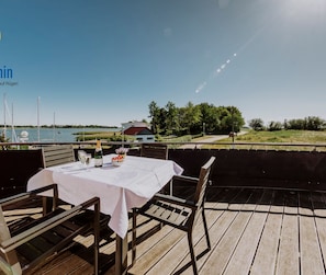 Eine Holzterrasse mit Tisch für zwei Personen, mit Blick auf ein Gewässer und ein Haus im Hintergrund.