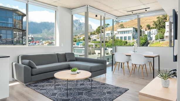 A modern living room with a grey sectional sofa, a round coffee table with a plant, a black and white patterned rug, and a dining area with white chairs and a wooden table. - The Quarter Apartments (Cape Town)