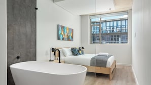 A modern bedroom with a large freestanding bathtub, a wooden bed, and a large window with a city view. - The Quarter Apartments (Cape Town)