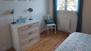 Une chambre avec une commode blanche, une petite table ornée de quelques objets décoratifs, une chaise et un lit recouvert d’une couvre-lit à motifs. Trois tableaux sont accrochés au mur et il y a une fenêtre avec des rideaux.