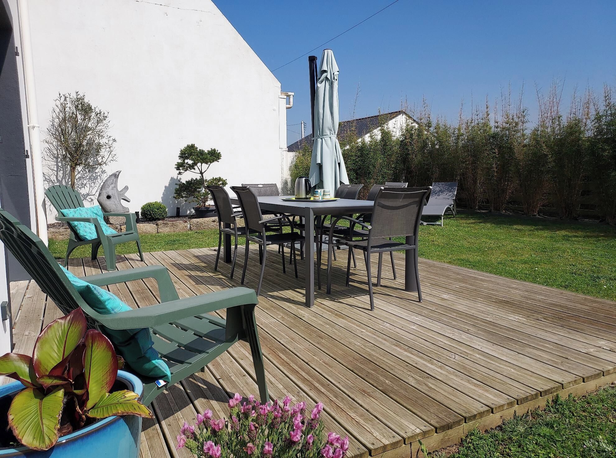 Une terrasse en bois, agréée de mobilier d’extérieur, notamment des chaises et une table, entourée de verdure et bordée d’un bâtiment blanc.