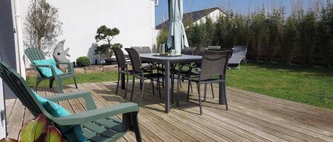 Une terrasse en bois, agréée de mobilier d’extérieur, notamment des chaises et une table, entourée de verdure et bordée d’un bâtiment blanc.
