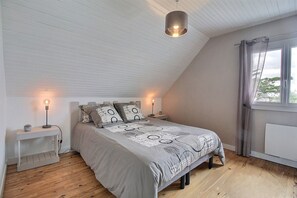Ein Schlafzimmer mit schrägen Wänden, einem Bett mit gemusterten Betttextilien, Nachttischlampen und einem Fenster mit Vorhängen.