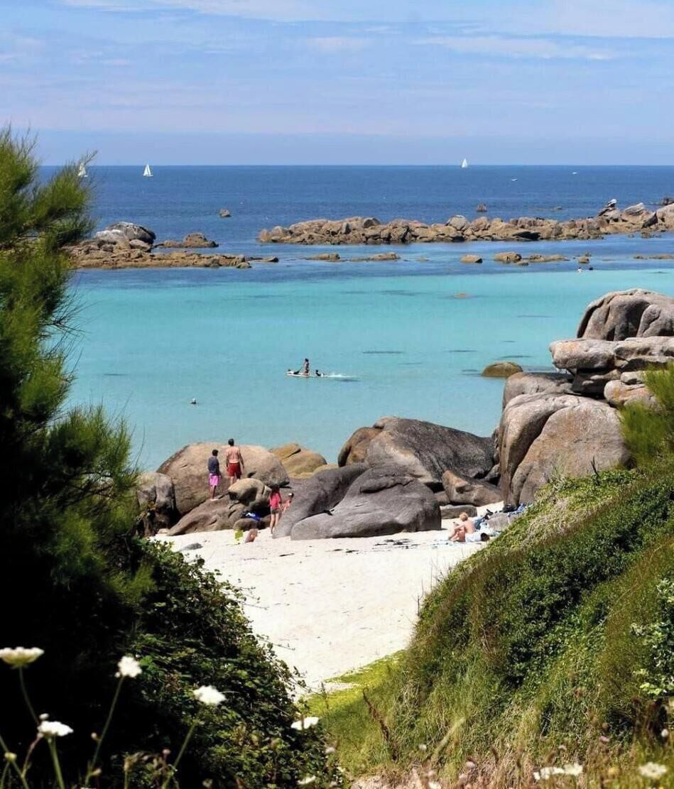 A beach with large rocks, clear turquoise water, and a distant sailboat.