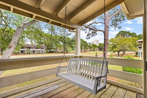 A wooden swing bench under a covered porch with a view of a park and buildings.
