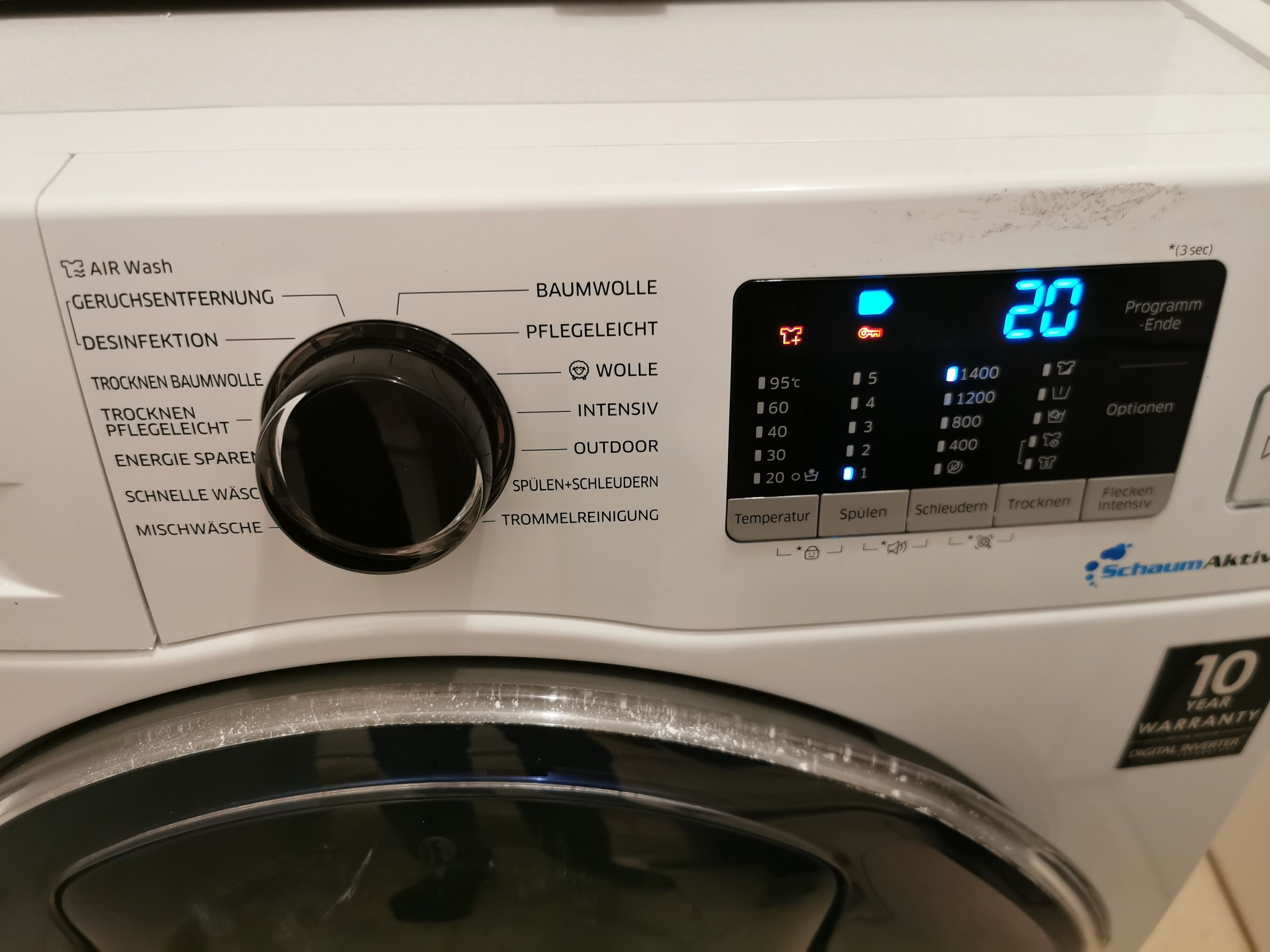 Een voor aanzicht van een wasmachine met een digitaal scherm, bedieningsknoppen en een overzicht van wasprogramma's.
