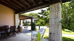 A covered patio area with a wooden table and chairs, a stone pillar, and a view of a swimming pool and greenery.