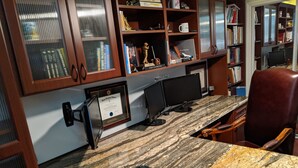 A home office with a granite countertop, a computer, and a chair. There are bookshelves with various items and a certificate on the wall.