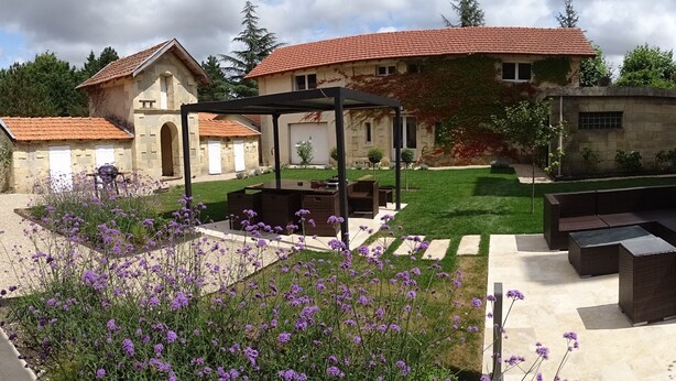 Un patio con una pérgola, mobiliario exterior y un edificio de piedra con techo de tejas rojas.