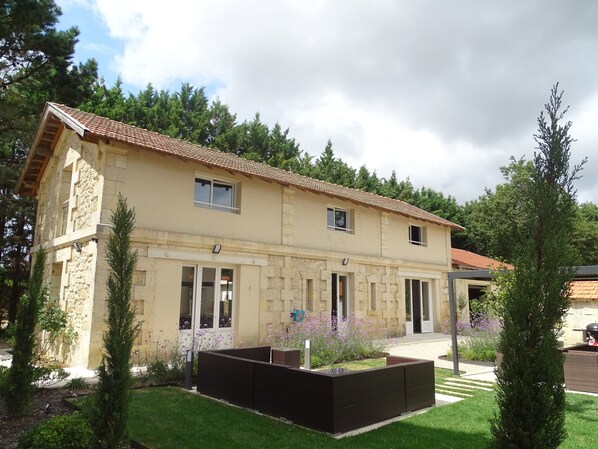 Un bâtiment en pierre de deux étages, avec un toit recouvert de tuiles, de nombreuses fenêtres et un jardin bien entretenu agrémenté d’arbustes et de fleurs.