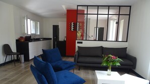 Una sala moderna con un sofá azul, un sofá gris y una mesa de centro blanca. Hay una zona de cocina con una pared de acento roja y una mesa de comedor con sillas.