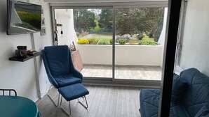 Un salon moderne avec un fauteuil bleu, une table turquoise et un canapé bleu. Un téléviseur est fixé au mur, et une porte-fenêtre coulissante donne accès à un balcon avec vue sur le jardin.