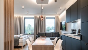 Une cuisine moderne avec des meubles de rangement de couleur sombre, une table à manger avec des chaises, un canapé et une grande fenêtre avec des rideaux.