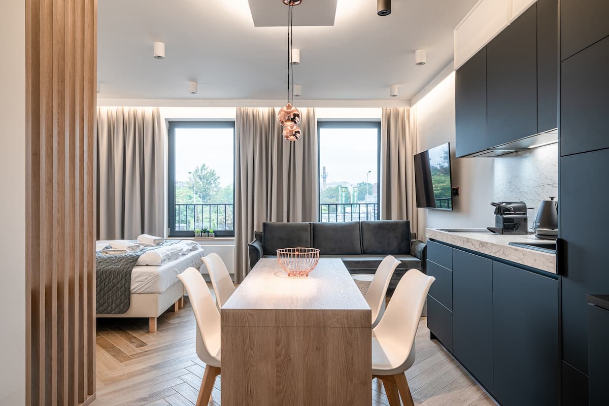 A modern kitchen with dark cabinets, a dining table with chairs, a sofa, and a large window with curtains.