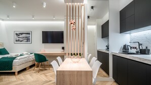 Une chambre d’hôtel moderne avec une table à manger en bois, des chaises blanches, un fauteuil vert, un lit recouvert d’une couverture verte, un téléviseur et un coin cuisine doté de meubles sombres et d’un plan de travail en marbre.