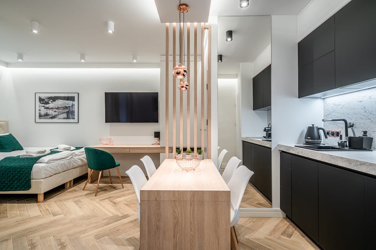 A modern hotel room with a wooden dining table, white chairs, a green chair, a bed with a green blanket, a TV, and a kitchen area with dark cabinets and a marble countertop.
