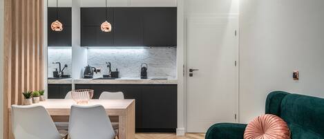 Cocina moderna con armarios oscuros, encimera de mármol e iluminación colgante. Hay un comedor con mesa de madera y sillas blancas. Se ve un sofá verde con un cojín rosa en primer plano.
