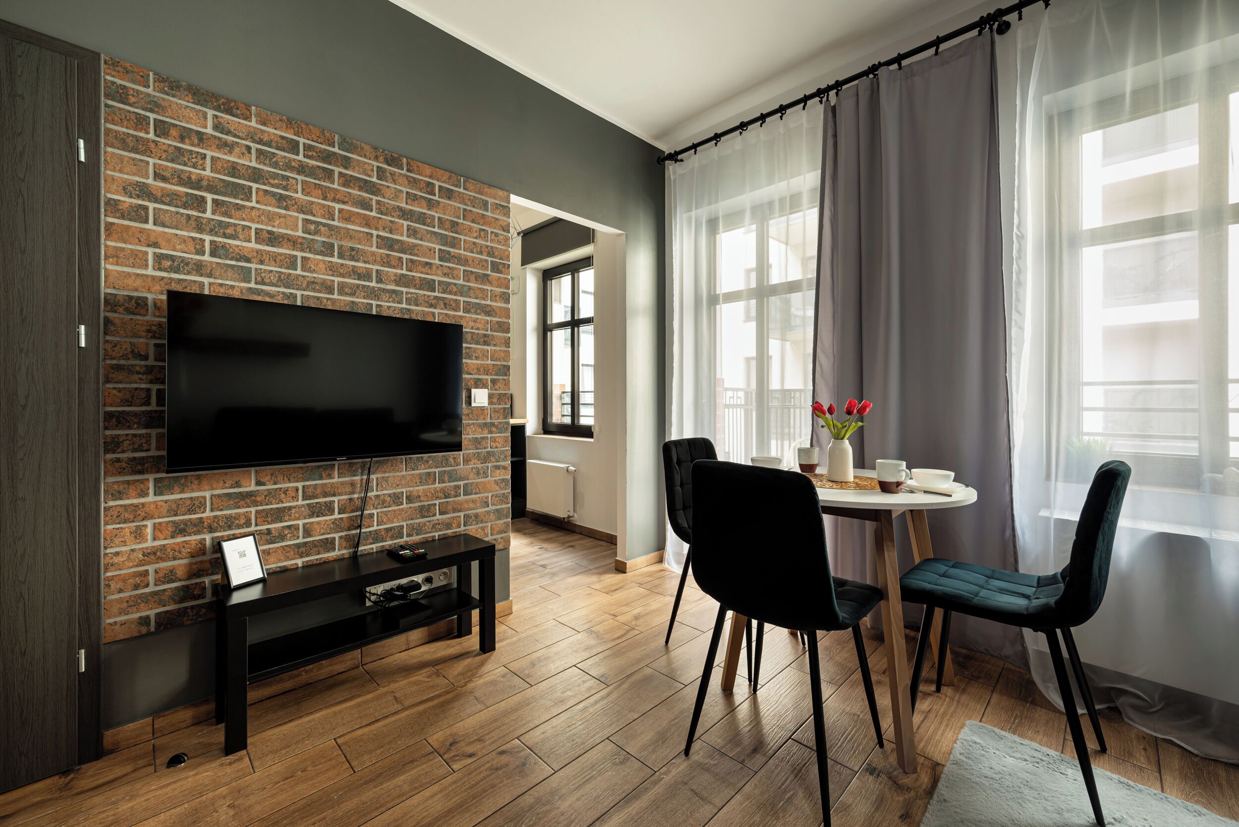 Una sala de estar con pared de ladrillo, un televisor de pantalla plana, una mesa de comedor con sillas y una ventana con cortinas de gasa.