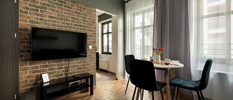 Una sala de estar con pared de ladrillo, un televisor de pantalla plana, una mesa de comedor con sillas y una ventana con cortinas de gasa.