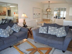 Ein Wohnzimmer mit zwei grauen Sofas, gemusterten Kissen, einem Glastisch, Holzboden, einem Essbereich mit weißem Tisch und Stühlen sowie einer durch eine Glasschiebetür sichtbaren Küche.