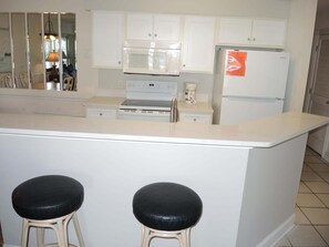 A kitchen with white cabinets, a microwave, a stove, a refrigerator, and two stools. - 1202m 1 Bedroom Condo by RedAwning (Calabash)