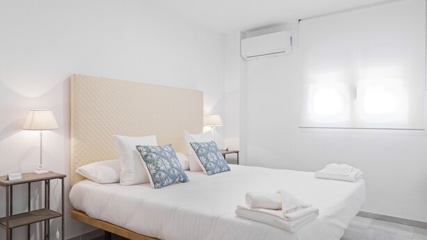 A neatly arranged hotel room with a large bed, two bedside tables with lamps, a wall-mounted air conditioner, and a window with blinds. - Pompidou 20 - 2 Rooms (Málaga)