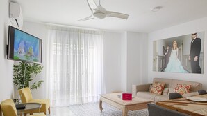 A modern living room with a ceiling fan, a flat-screen TV, a large window with sheer curtains, a sofa with patterned cushions, a coffee table, and a dining table with chairs. - Pompidou 20 - 2 Rooms (Málaga)
