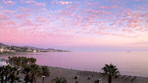 A beach with palm trees and umbrellas, a calm sea, and a cityscape in the background. - Malagueta - Miradore II (Málaga)
