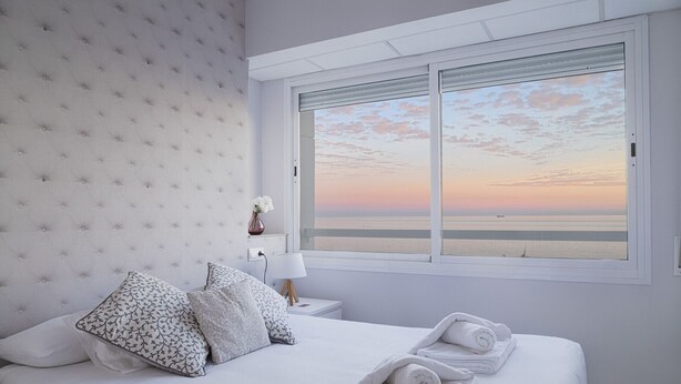 A bedroom with a large bed, a nightstand, a lamp, and a window with a sunset view. - Malagueta - Miradore II (Málaga)