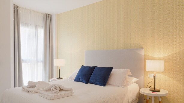 Ein Hotelzimmer mit einem großen Bett, zwei Nachttischen mit Lampen, einem zusammengerollten Handtuch auf dem Bett und einem Fenster mit Vorhängen.