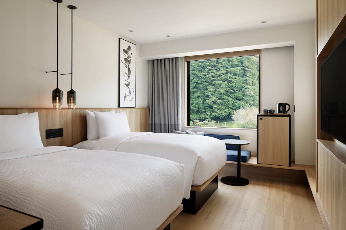 Une chambre d’hôtel moderne avec deux lits, une grande fenêtre donnant sur la forêt, une petite table et un téléviseur.