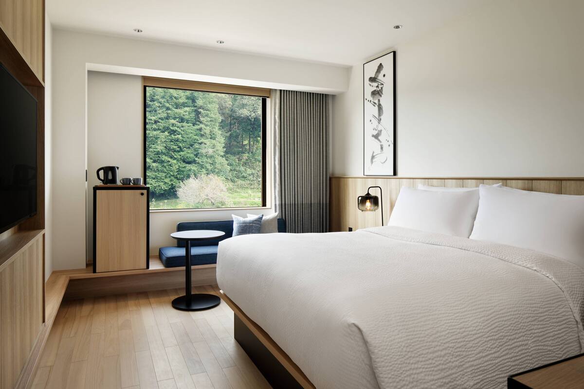Une chambre d’hôtel moderne dotée d’un grand lit, d’un petit coin salon, d’une fenêtre donnant sur les arbres et d’un téléviseur à écran plat.