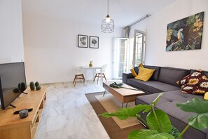 Un salon moderne avec un canapé gris, une table basse en bois, un téléviseur et un coin repas comprenant une table et des chaises blanches.