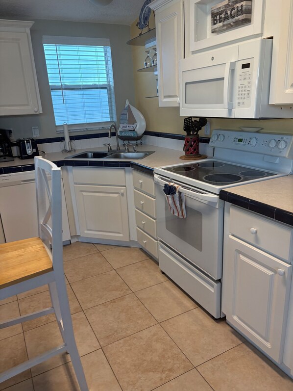 A kitchen with white cabinets, a sink, a stove, and a window with blinds. - Island Therapy (Cudjoe Key)