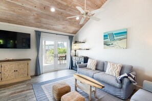 A living room with a grey sofa, a wooden bench, a TV, a ceiling fan, and a painting of a beach scene.