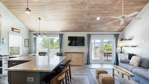 Una cocina moderna con una isla grande, taburetes de bar, una zona de comedor con mesa y sillas, un ventilador de techo y un televisor montado en la pared.